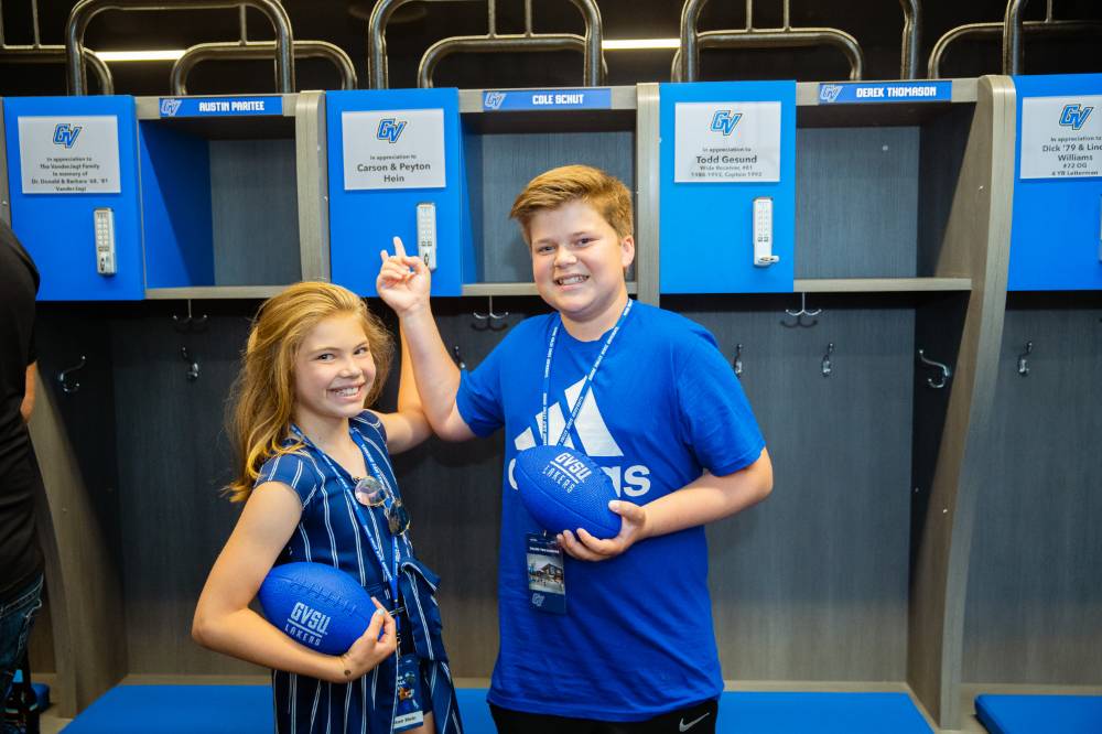 Two children holding footballs, pointing to a named locker at the Jamie Hosford Football Center dedication.